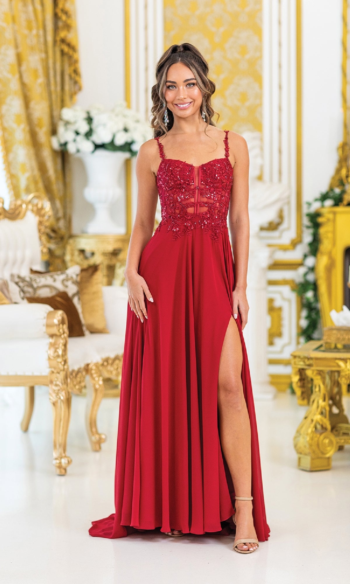 A woman stands indoors in the Lace-Corset Long Prom Dress: Dancing Queen 4531—a red, sleeveless gown with a thigh-high slit—surrounded by elegant floral arrangements and gold and white furniture.